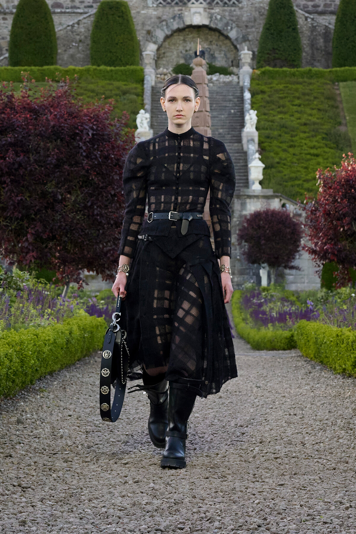 Model in a black sheer lace dress with a wide belt and knee-high boots walks along a gravel garden path, carrying a studded black bag.
