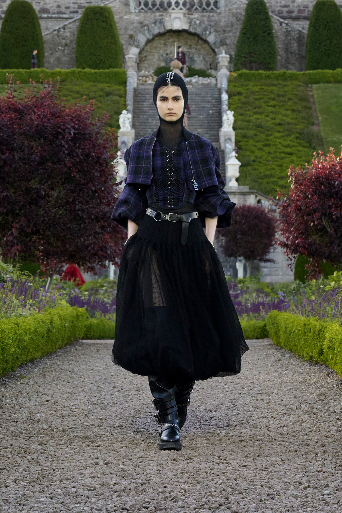 Model in a gothic outfit with corset, plaid shawl, and black tulle skirt walks toward the camera in a formal garden with stone steps in the background.