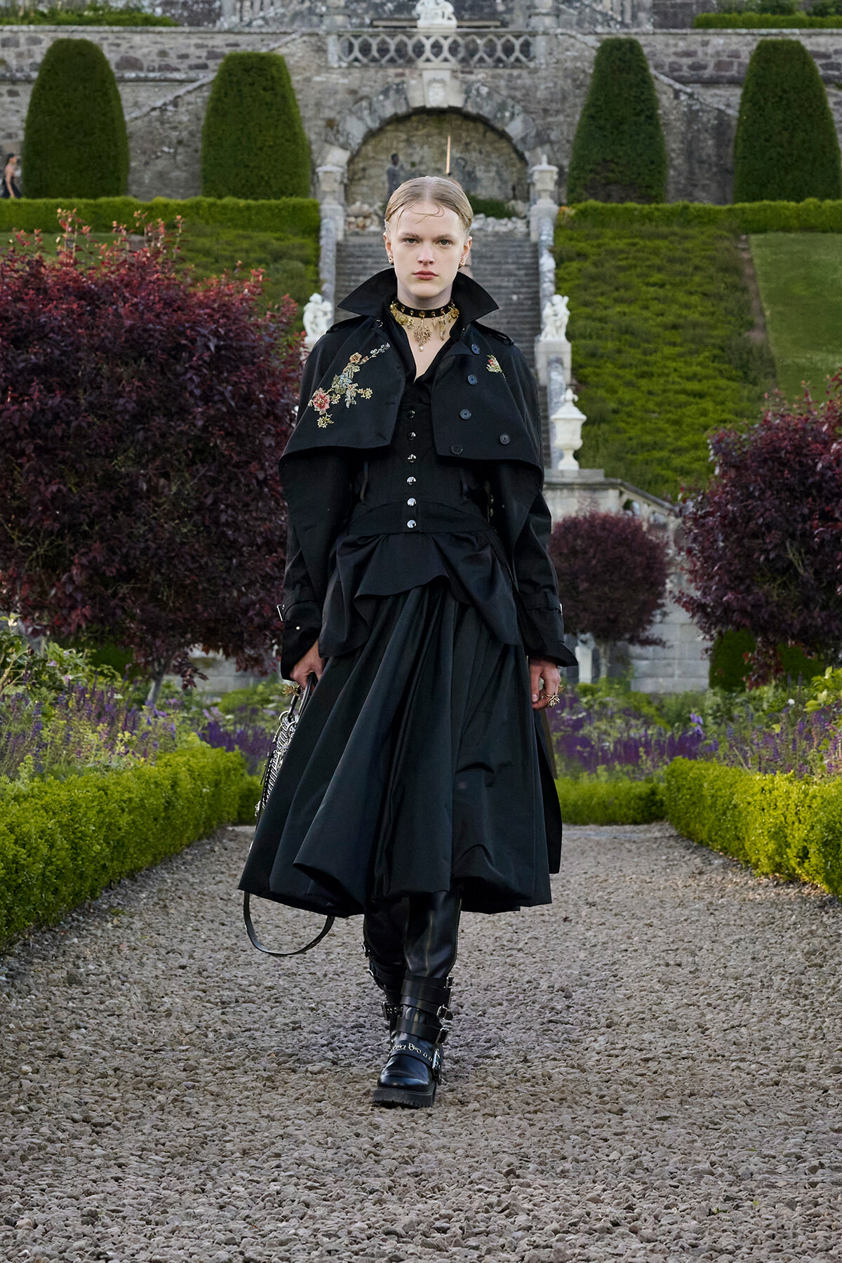 Model walking a gravel garden path in a black Victorian-inspired outfit with an embroidered cape and choker