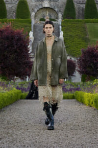 Model walking toward the camera on a gravel garden path, wearing an oversized olive-green jacket over a beige lace dress and black boots, with a formal garden backdrop.