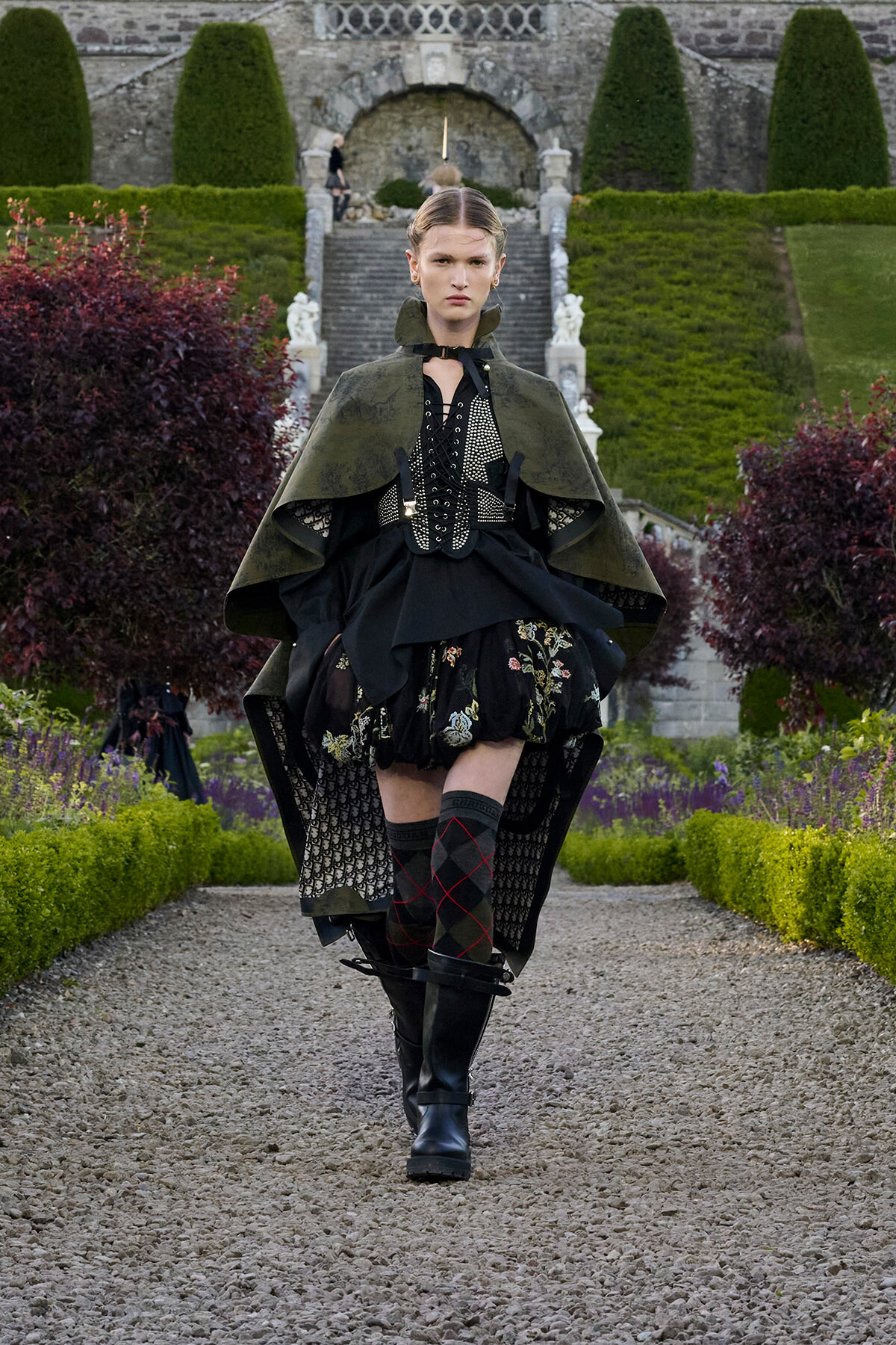 Model walking down a gravel garden path in a dramatic green cape and black Victorian-inspired outfit with boots.