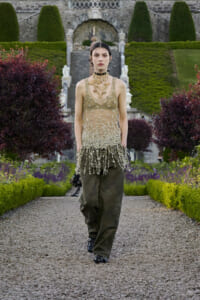 Model walks down a gravel garden path in a beaded sheer top, layered gold necklaces, and olive wide-leg pants.