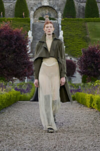 Model walks along a gravel garden path in an olive trench coat over a cream lace top and a sheer beige mesh skirt on a formal garden runway.
