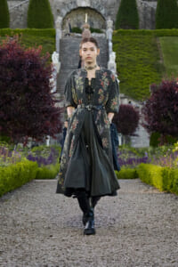 Model walking toward the camera in a dark floral long coat, corset, and black boots along a gravel garden path with steps behind.