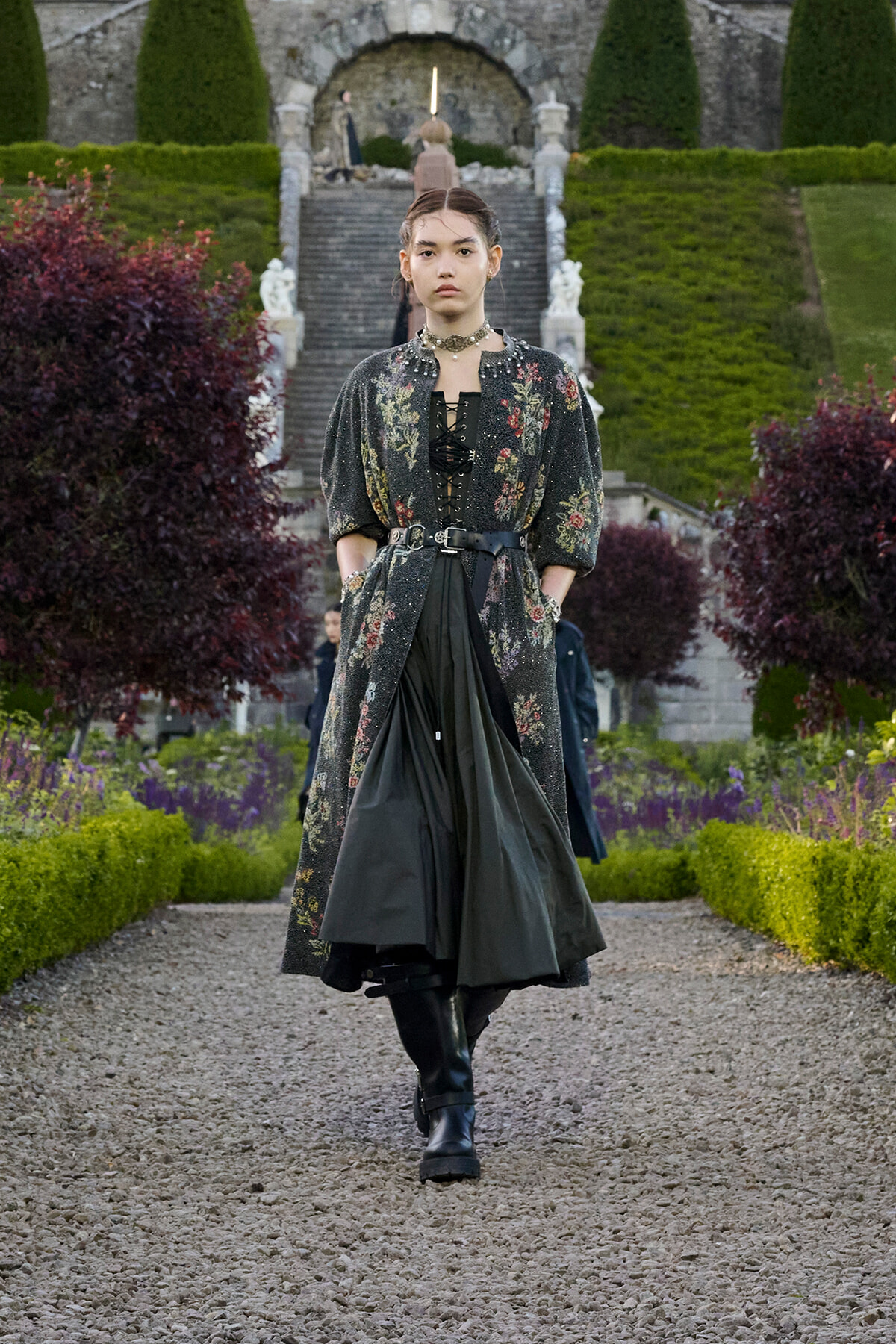 Model walking toward the camera in a dark floral long coat, corset, and black boots along a gravel garden path with steps behind.
