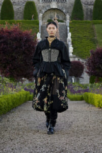 Model wearing a black peplum jacket with a beaded corset belt and a floral-embroidered skirt walks along a gravel garden path.