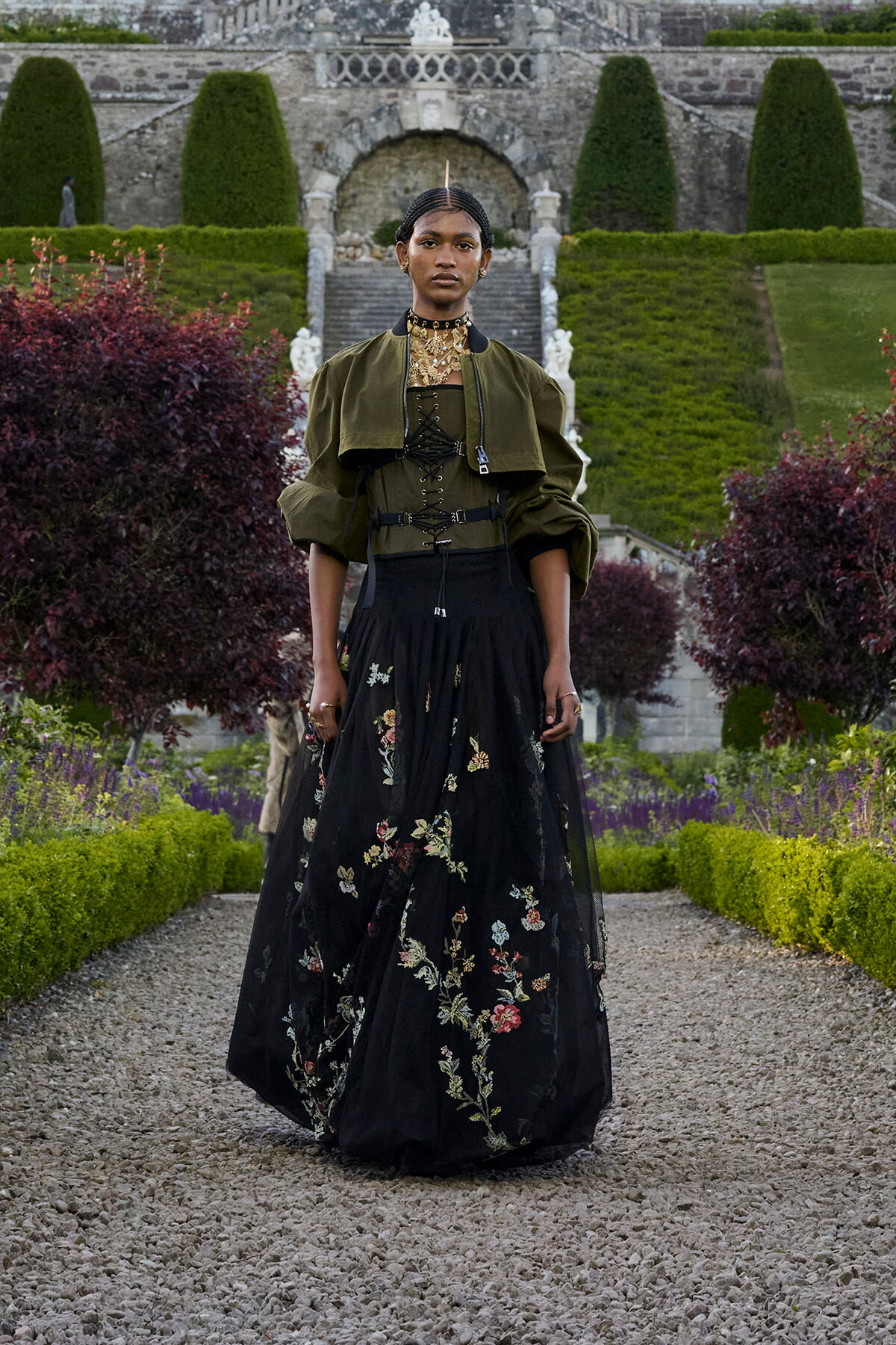 Person in a black floral-embroidered gown with an olive-green cropped jacket and gold choker, standing in a formal garden.