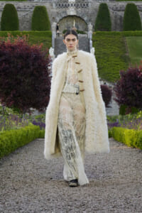 Model in a cream boucle coat over a lace dress walks along a gravel garden path with formal stone steps and trimmed hedges in the background.