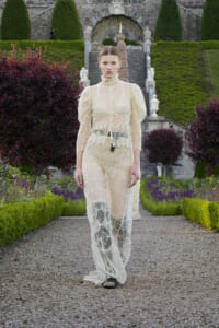 Model walking toward the camera on a gravel garden path in a cream lace outfit with a high collar.