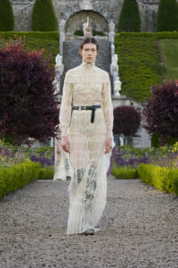 Model walking a gravel path in a formal cream lace gown with high neck and black belt, in a manicured garden setting.