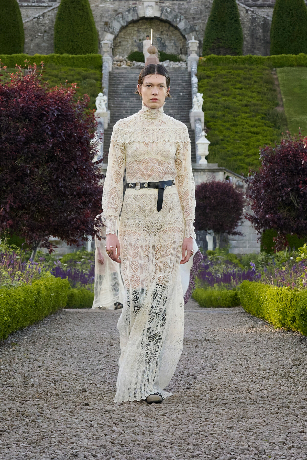 Model walking a gravel path in a formal cream lace gown with high neck and black belt, in a manicured garden setting.
