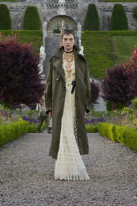 Model wearing a cream lace gown under a long olive trench coat stands on a gravel garden path with manicured hedges and stone arches in the background.