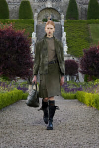 Model walking down a gravel garden path in an olive-green trench coat, black knee-high boots, and a quilted handbag, with a formal stone staircase and manicured hedges in the background.
