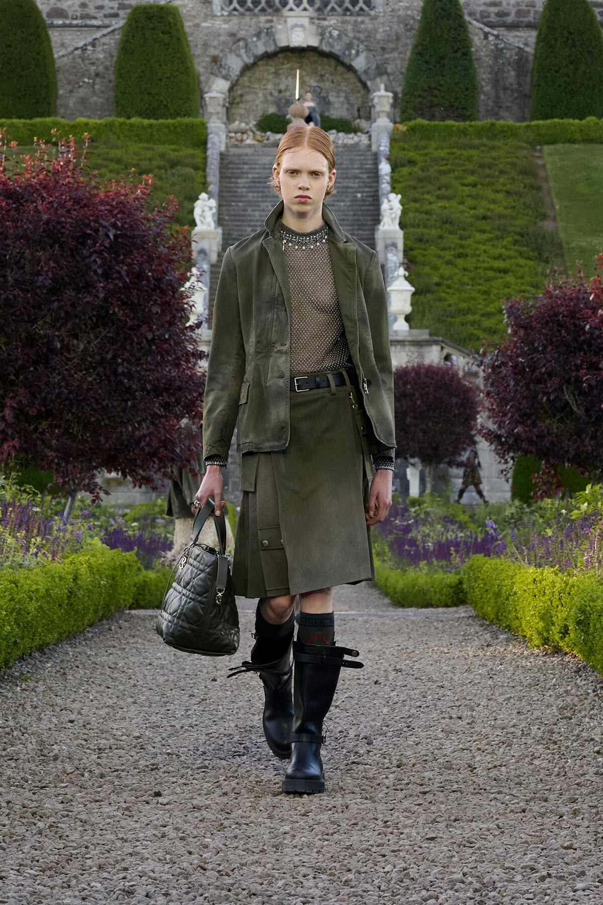 Model walking down a gravel garden path in an olive-green trench coat, black knee-high boots, and a quilted handbag, with a formal stone staircase and manicured hedges in the background.
