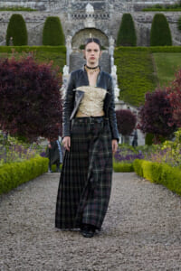 A woman wearing a black leather jacket, beige lace top, and long plaid skirt walks along a gravel garden path with trimmed hedges and a stone staircase in the background.