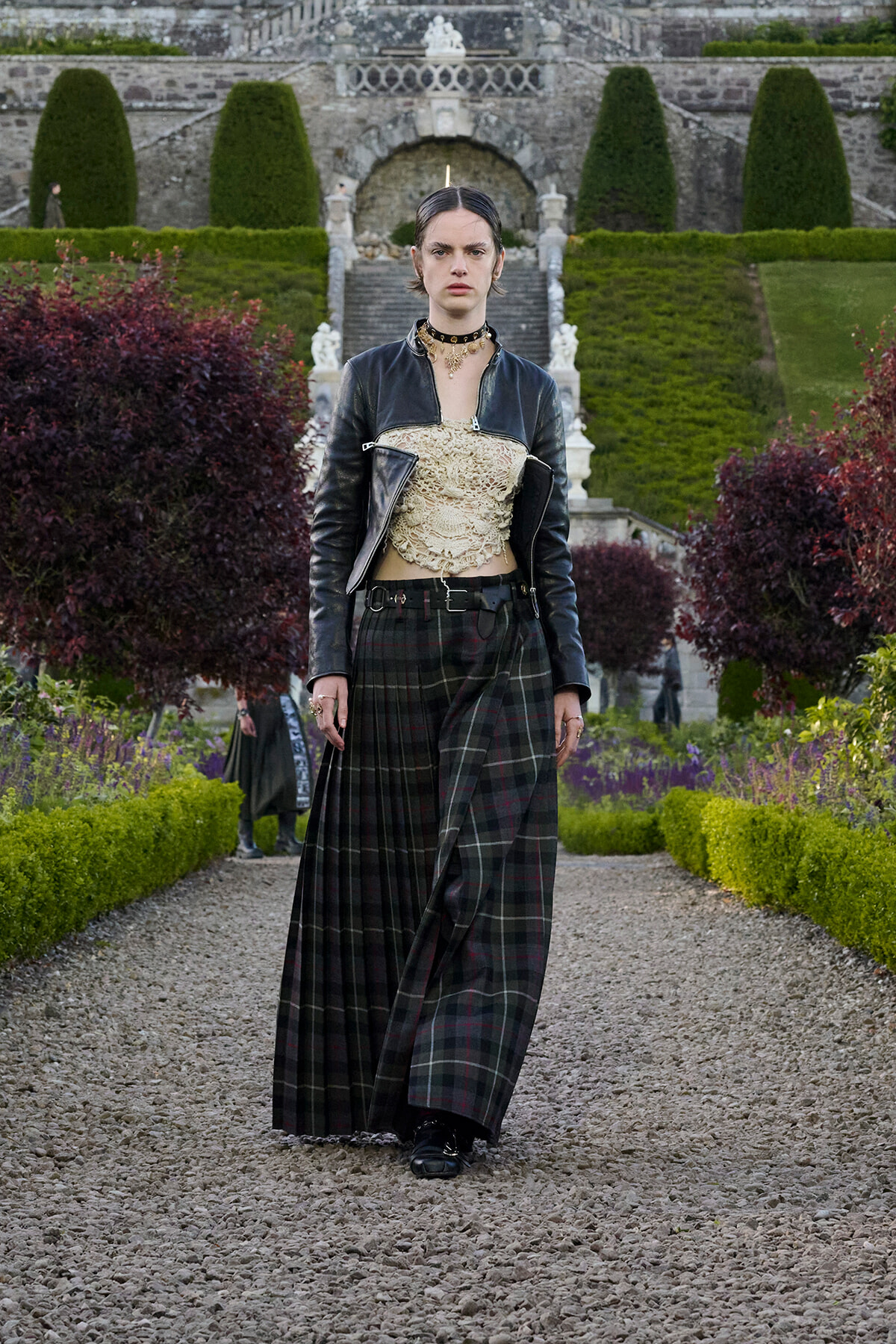 A woman wearing a black leather jacket, beige lace top, and long plaid skirt walks along a gravel garden path with trimmed hedges and a stone staircase in the background.
