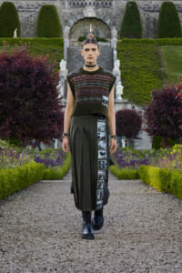 Model walking toward camera in a formal garden, wearing a dark green knitted sleeveless sweater over a long pleated skirt with a vertical strip of black-and-white photos, black boots, and a choker, with manicured hedges and stone steps in the background.