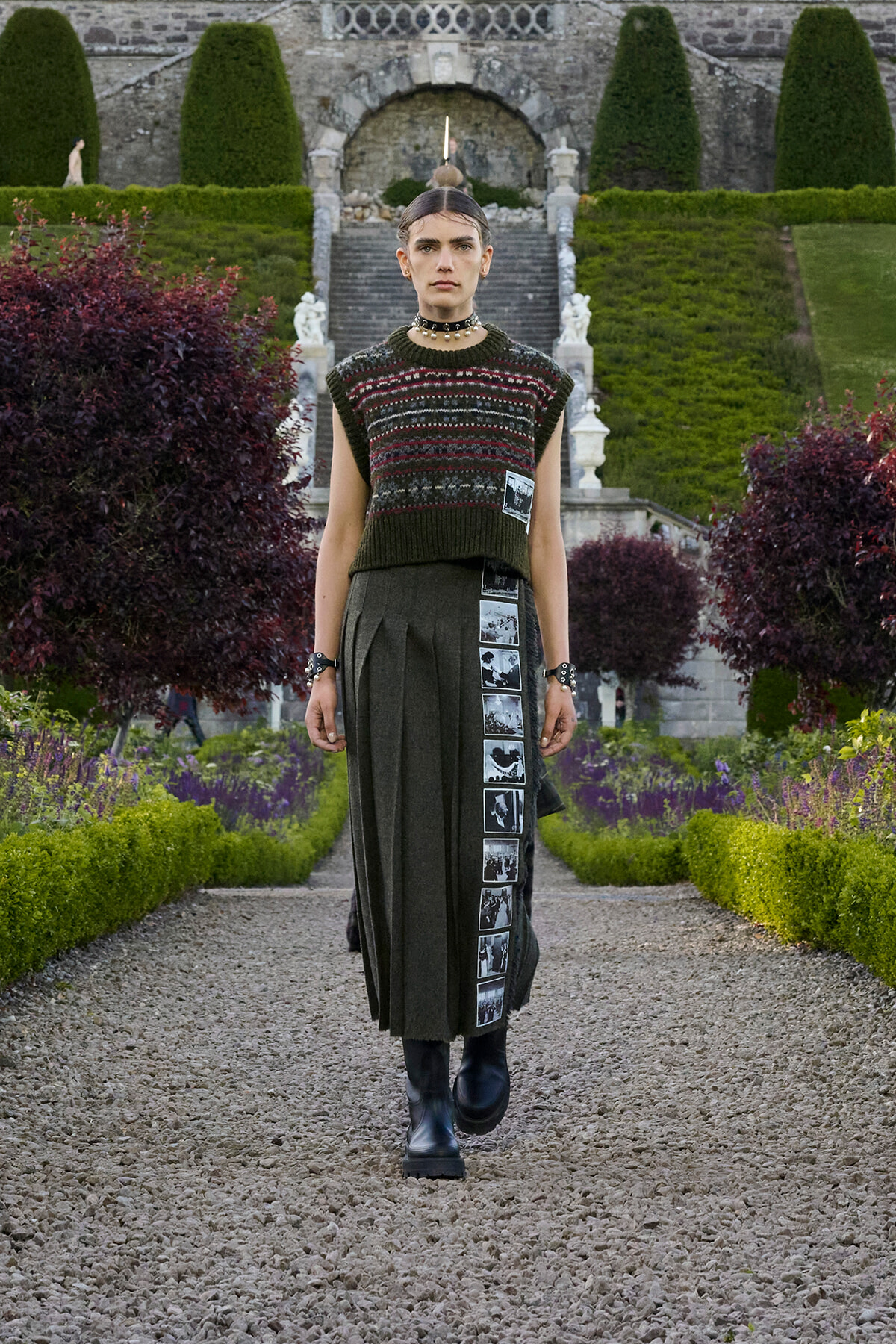 Model walking toward camera in a formal garden, wearing a dark green knitted sleeveless sweater over a long pleated skirt with a vertical strip of black-and-white photos, black boots, and a choker, with manicured hedges and stone steps in the background.