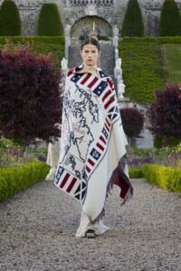 Model standing on a gravel path in a formal garden, wrapped in a white blanket with red‑white stripes and blue flag motif, fringed at the edge, with stone steps and manicured hedges in the background.