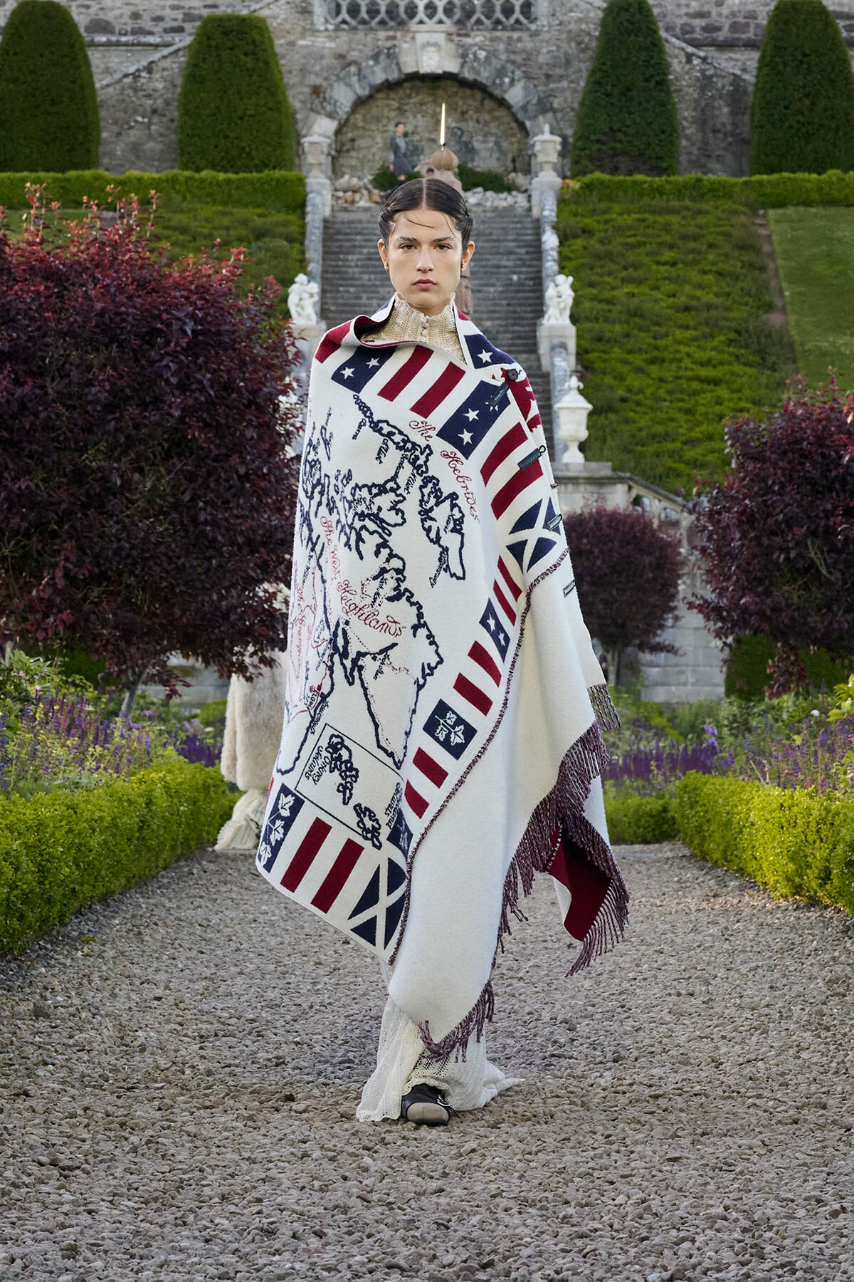 Model standing on a gravel path in a formal garden, wrapped in a white blanket with red‑white stripes and blue flag motif, fringed at the edge, with stone steps and manicured hedges in the background.