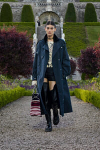Model walking toward camera in a navy trench coat, black boots, and a striped tote bag in a formal garden setting.
