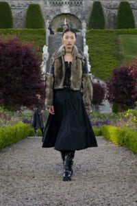 Fashion model walking down a gravel garden path in a fur-trimmed vest, black corset and long skirt, black boots.