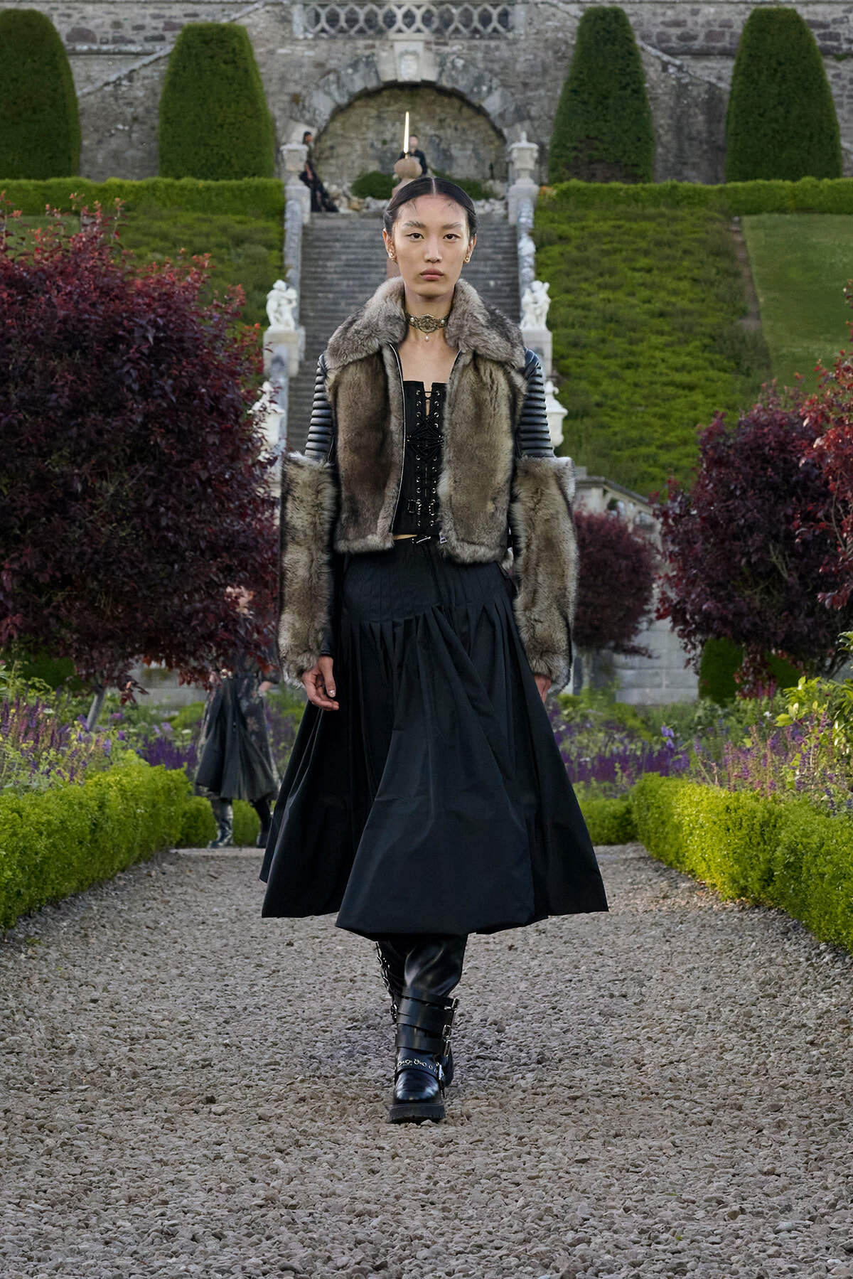 Fashion model walking down a gravel garden path in a fur-trimmed vest, black corset and long skirt, black boots.