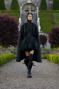 Full-length fashion model in an all-black avant-garde outfit walking on a gravel path with a formal garden and stone stairs behind her.