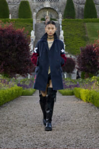 Person walking toward the camera in a navy coat with red sleeves and white stars, paired with black knee-high boots, in a formal garden path.