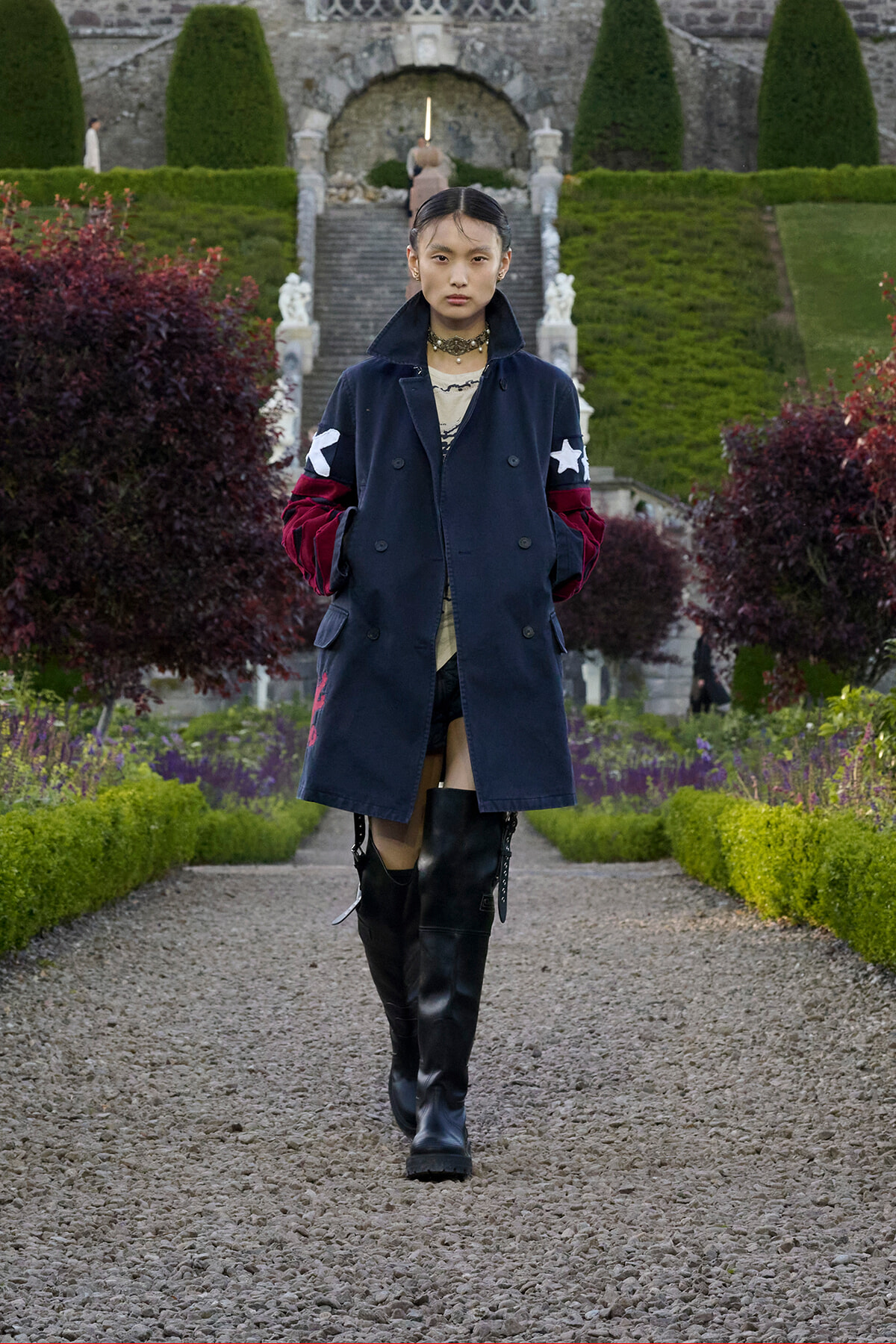 Person walking toward the camera in a navy coat with red sleeves and white stars, paired with black knee-high boots, in a formal garden path.