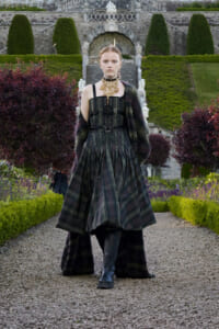 Model walks down a gravel courtyard in a dark plaid ball gown with corset bodice and fur stole, set in a formal garden backdrop.