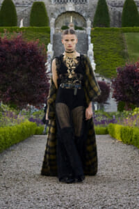 Model in a black lace jumpsuit and striped fur coat walks along a gravel garden path with trimmed hedges.