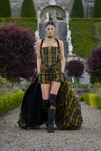 Model walking a gravel path in a green‑and‑black plaid corset dress with a long cape in a formal garden setting.