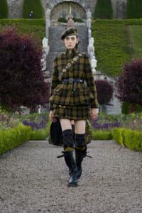 Woman in a brown and black plaid military-style outfit walking on a gravel garden path, with a crossbody belt and tall black boots near a formal stone staircase.