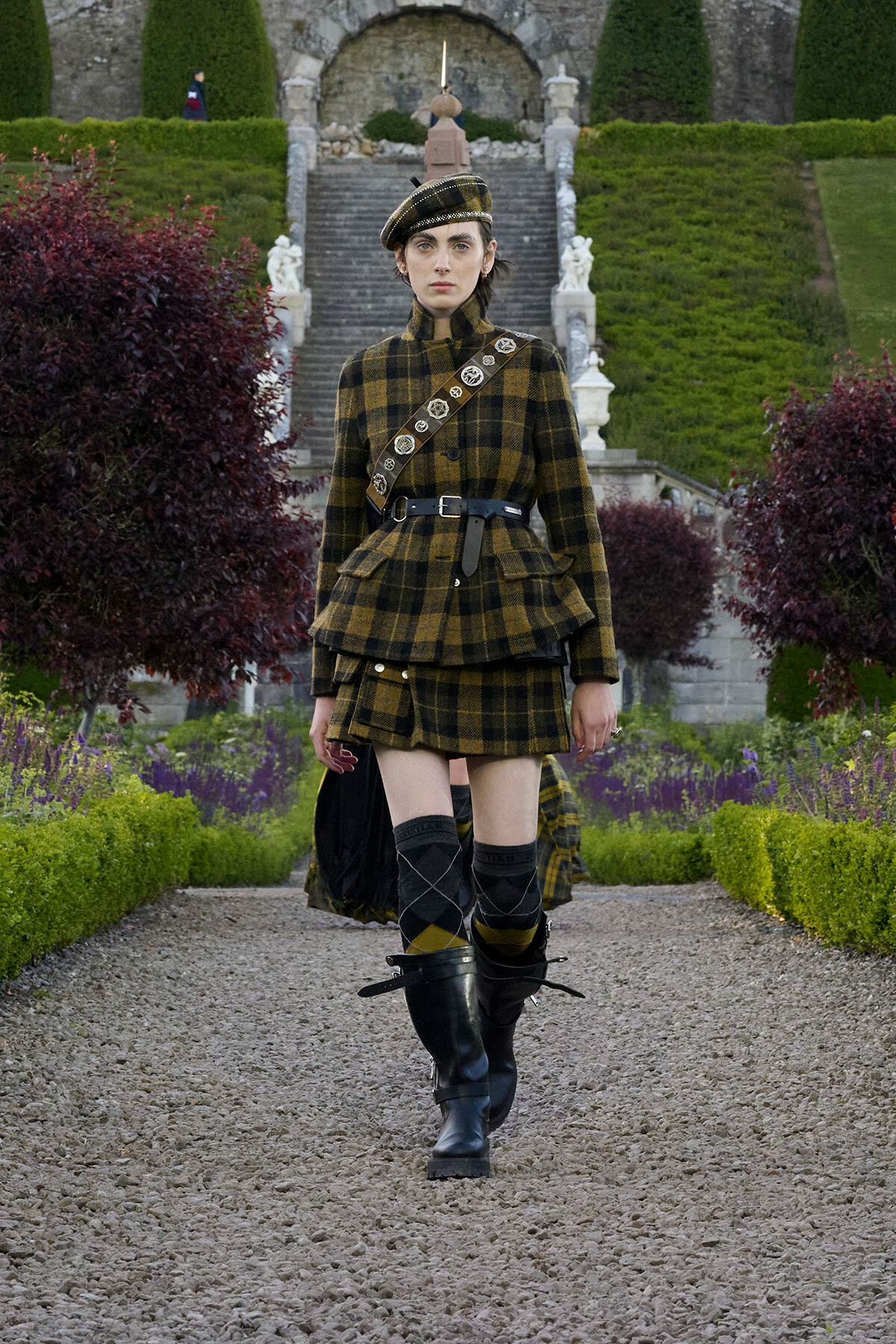 Woman in a brown and black plaid military-style outfit walking on a gravel garden path, with a crossbody belt and tall black boots near a formal stone staircase.