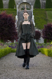 Model in a black corset dress with sheer dotted sleeves, walking on a gravel path in a formal garden with manicured hedges and stairs behind her.