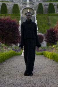 Person walking toward camera wearing a long black knit outfit in a formal garden with gravel path and trimmed hedges; stone stairs in the background.