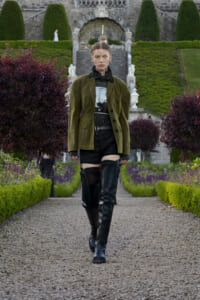 Model walks a gravel garden runway in an olive-green blazer, black shorts, belt, and tall black boots with a sheer face veil.