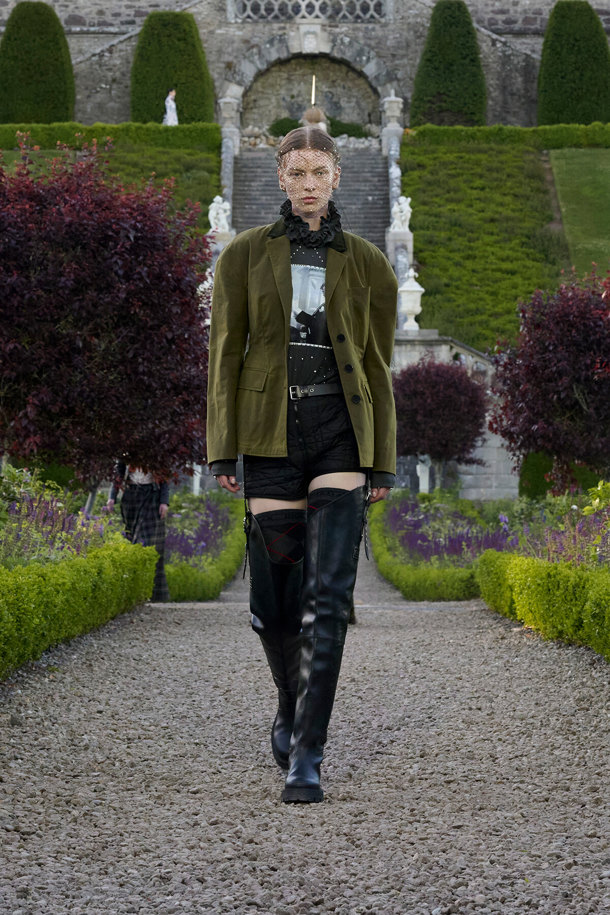 Model walks a gravel garden runway in an olive-green blazer, black shorts, belt, and tall black boots with a sheer face veil.