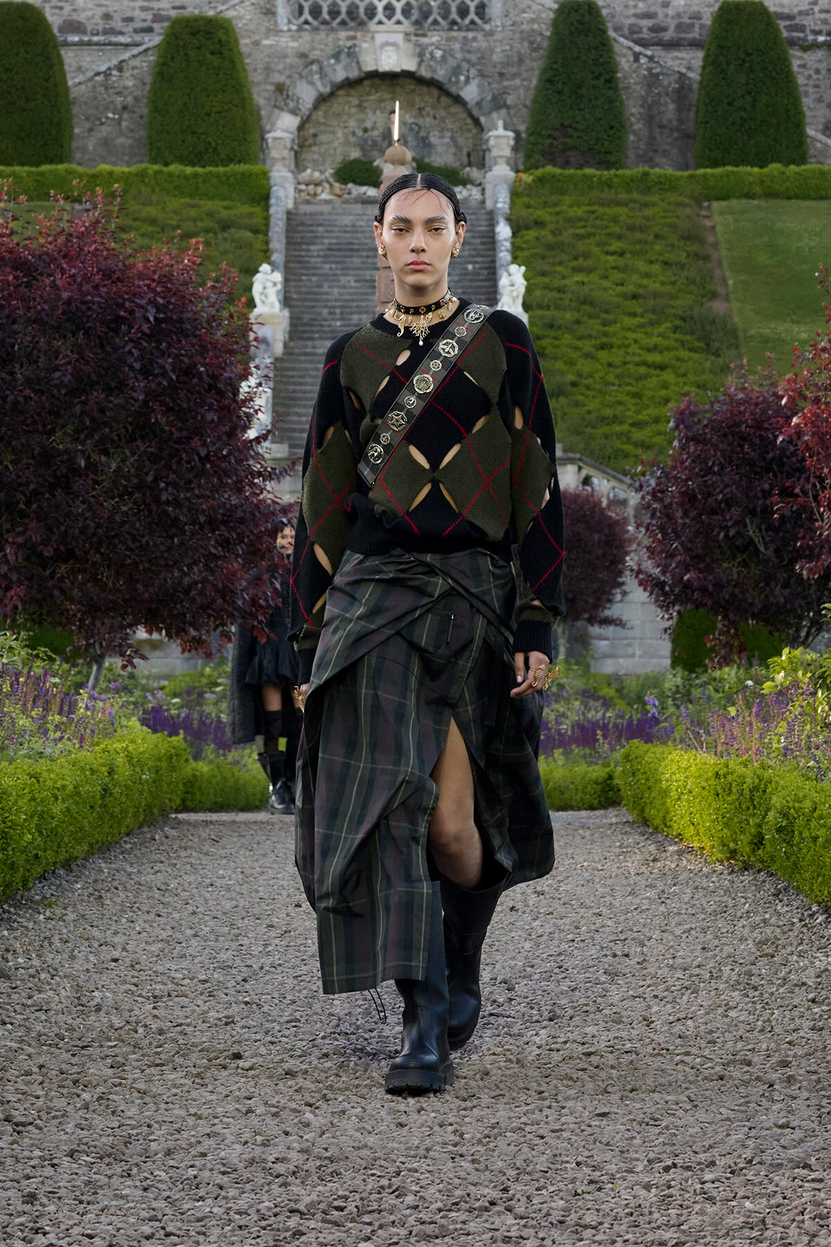 Model walking down a gravel garden path in a dark argyle sweater, plaid wrap skirt, and boots, with formal stone stairs behind.