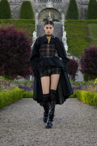 Model walking down a gravel path in a dark Victorian-inspired outfit with a corset and high-low cape, in a formal garden backdrop.