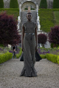 Model in a metallic chainmail gown with a black belt walks down a gravel garden path, stone stairs and manicured hedges in the background.