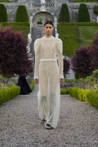 Model walking a gravel garden path in an ivory lace jumpsuit with high neck and puff sleeves, set against formal stone gardens behind.