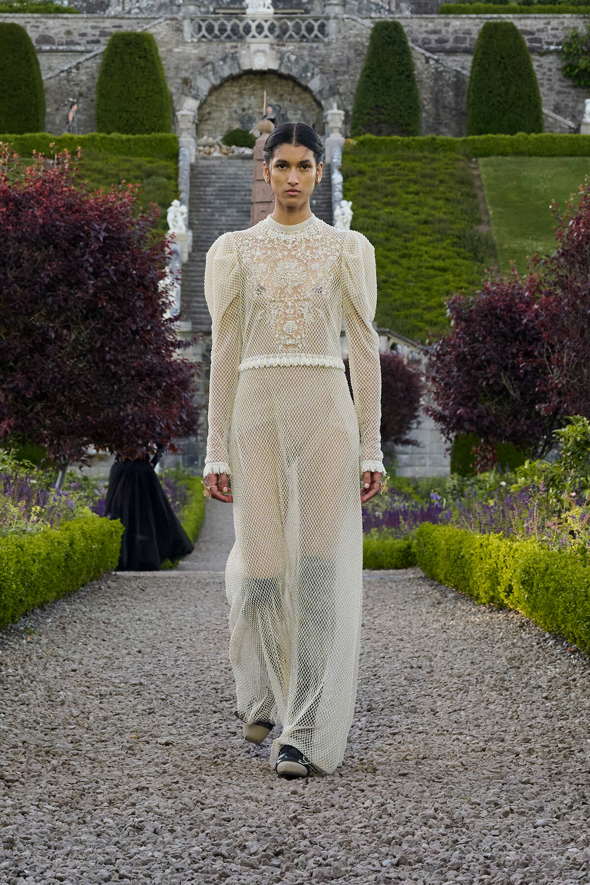 Model walking a gravel garden path in an ivory lace jumpsuit with high neck and puff sleeves, set against formal stone gardens behind.