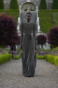 Model in a long metallic dress with puffed shoulders and a belt, standing on a gravel path in a formal garden.