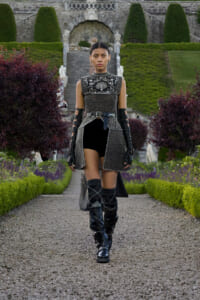 Model walking down a gravel path in a formal, metallic-themed outfit with black and silver armor-like top, belt, and knee-high boots in a manicured garden setting with stone stairs behind her.