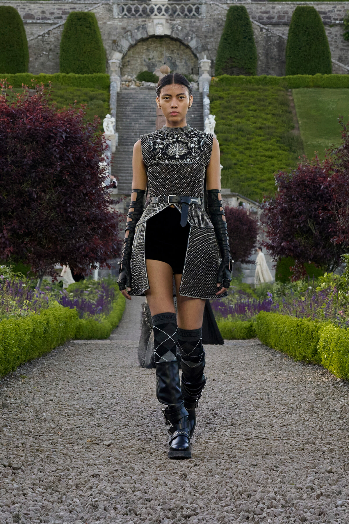 Model walking down a gravel path in a formal, metallic-themed outfit with black and silver armor-like top, belt, and knee-high boots in a manicured garden setting with stone stairs behind her.