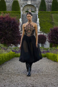 Model in a black embroidered halter gown with a sheer skirt walking along a gravel garden path, formal garden in background] }{