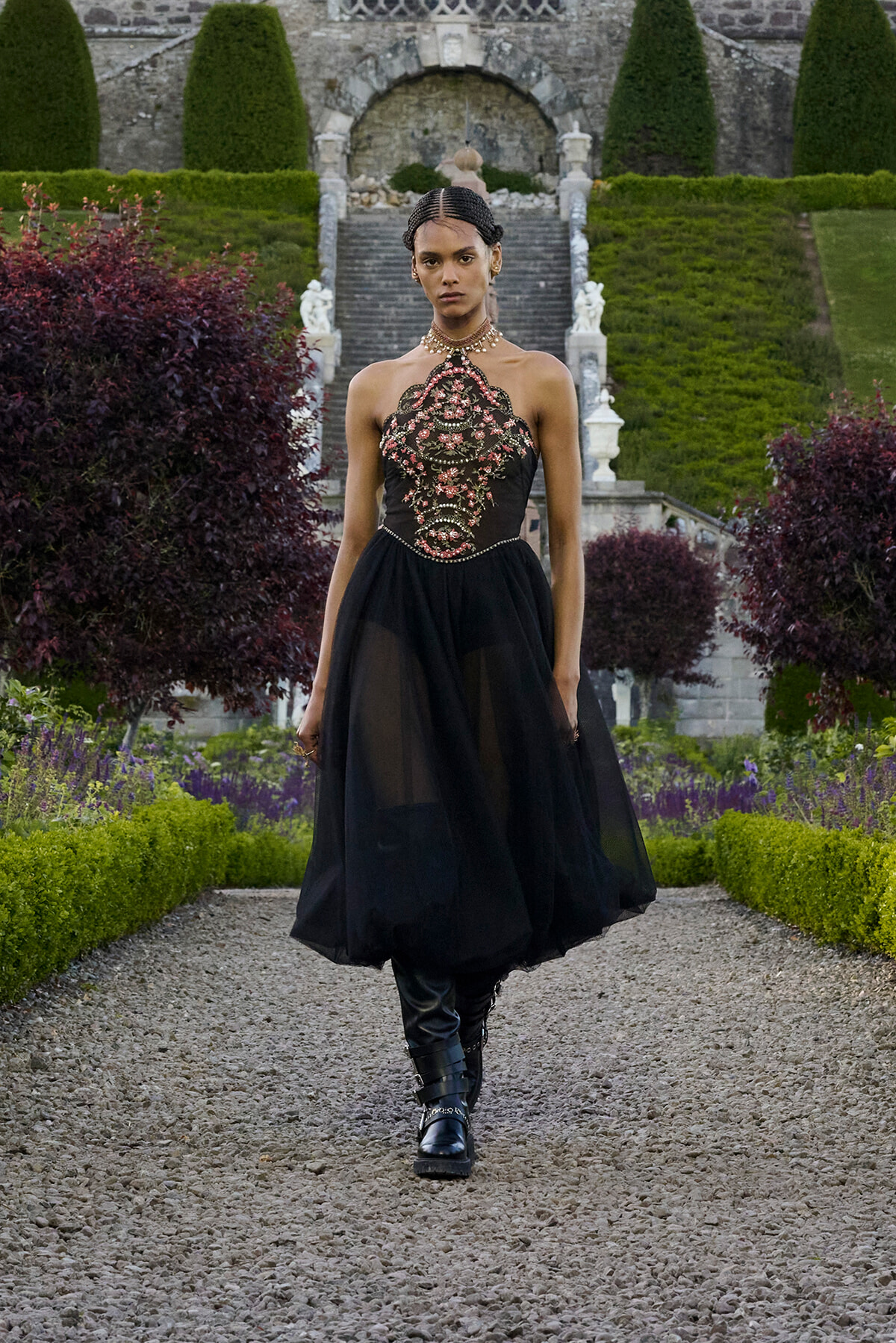 Model in a black embroidered halter gown with a sheer skirt walking along a gravel garden path, formal garden in background] }{