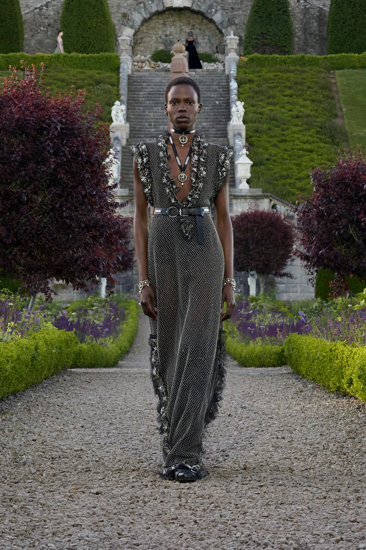 Female model walking down a gravel path in a metallic embellished gown with a belt, jewelry, and a formal garden backdrop with stone stairs and sculptures.